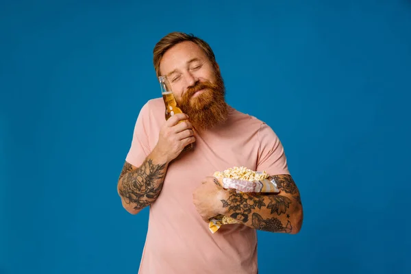 Pleased ginger bearded man holding beer and popcorn while standing isolated over blue studio background