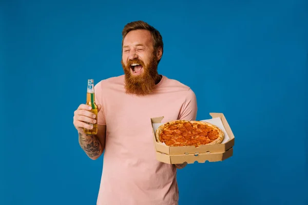 Portrait of excited ginger bearded man screaming while holding beer and pizza isolated over blue studio background