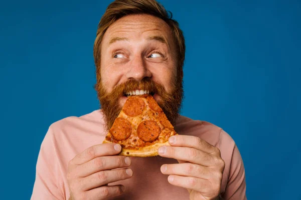 Bearded happy man eating pizza while standing isolated over blue background