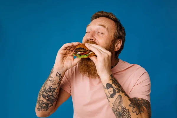 Bearded hungry man eating burger while standing isolated over blue background