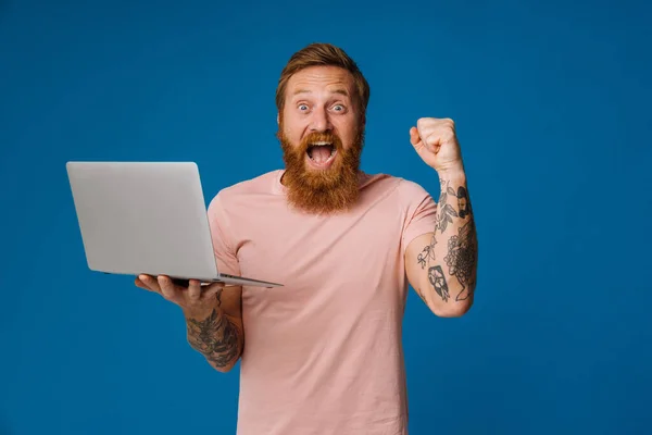 满脸胡须的快乐男人一边尖叫一边举着笔记本电脑 孤零零地站在蓝色的背景下 — 图库照片