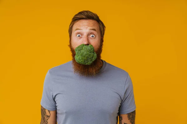 Bearded happy man eating broccoli while standing isolated over yellow background