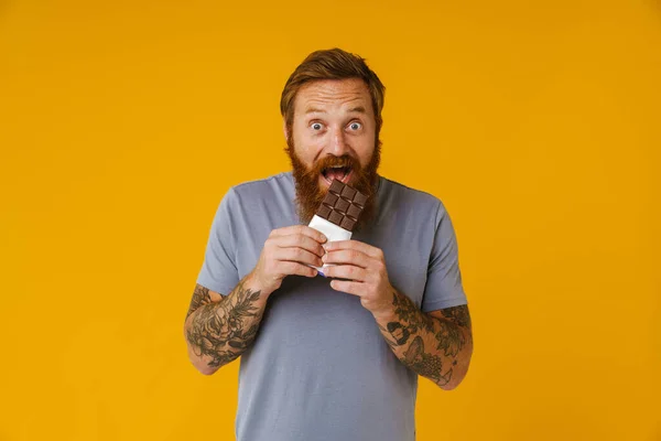 Bearded happy man eating chocolate bar while standing isolated over yellow background