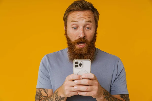 Bearded white man holding cellphone while standing isolated over yellow background