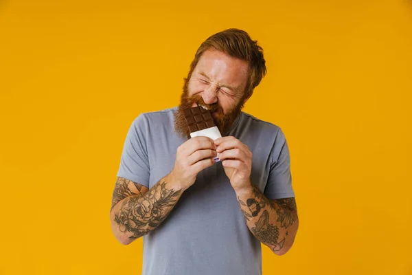 Bearded happy man eating chocolate bar while standing isolated over yellow background