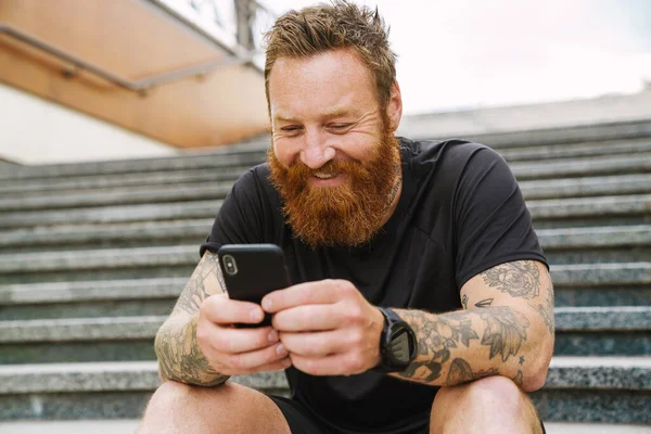 stock image Young handsome bearded tattooed redhead smiling man holding and using his phone, while sitting on the stairs outdoors