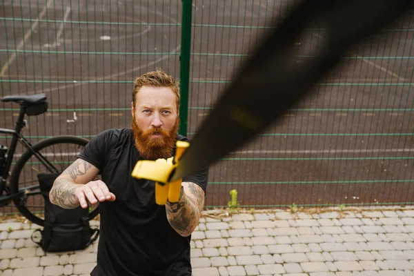 stock image Young handsome tattooed bearded serious redhead man looking up and doing exercises with TRX near bicycle at outdoor sports field