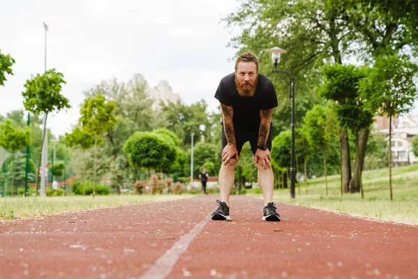 stock image Portrait of adult bearded redhead man leaning on his knees and looking at camera, while standing on running track outdoors