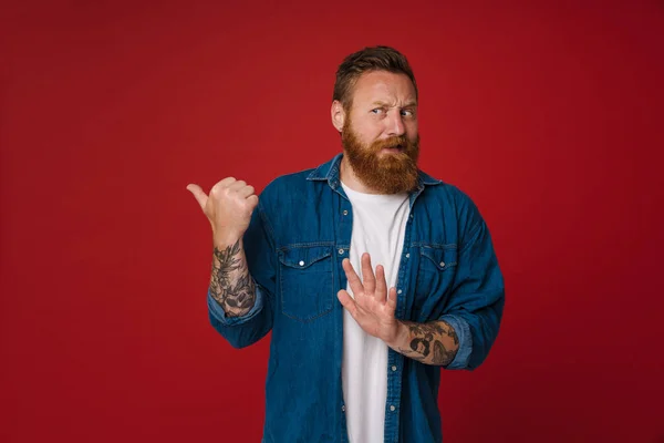 大胡子纹身的英俊的红头发男人用大姆指着旁边 做着拒绝的手势 把目光投向一边 站在孤立的红色背景上 — 图库照片