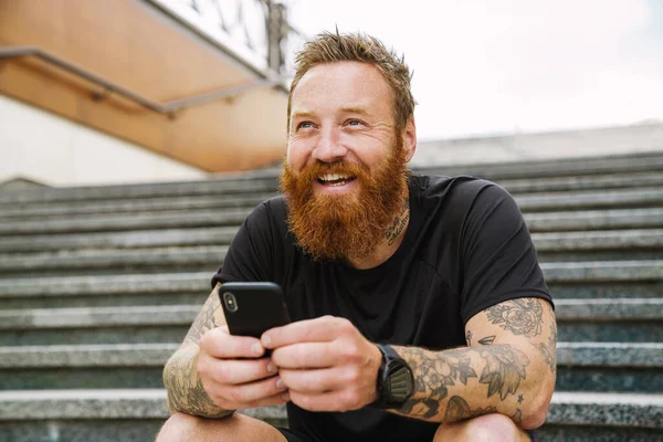 Ginger bearded sportsman using mobile phone while sitting on stairs outdoors