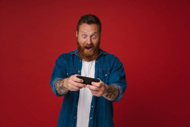 Ginger heyecanlı adam pembe arka planda izole bir şekilde cep telefonuyla oyun oynuyor.