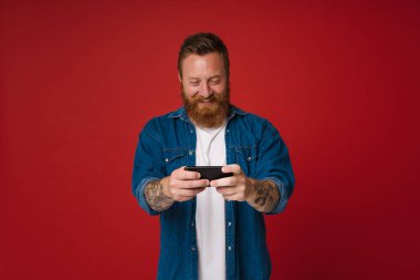 Ginger heyecanlı adam pembe arka planda izole bir şekilde cep telefonuyla oyun oynuyor.