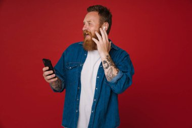Ginger kulaklıkta müzik dinleyen ve kırmızı arka planda izole edilmiş cep telefonu kullanan heyecanlı bir adam.