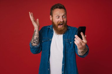 Kızıl, kot pantolon giyen, cep telefonu kullanan ve kırmızı arka planda izole edilmiş hareketler yapan heyecanlı bir adam.