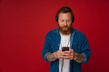 Kızıl saçlı, kulaklıklı, müzik dinleyen ve kırmızı arka planda izole edilmiş bir cep telefonu kullanan beyaz bir adam.