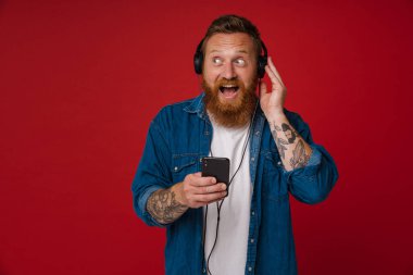 Kızıl saçlı, kulaklıklı, müzik dinleyen ve kırmızı arka planda izole edilmiş bir cep telefonu kullanan sürpriz bir adam.