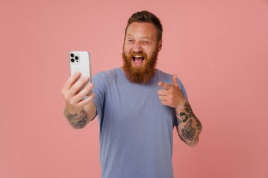 Kızıl beyaz adam cep telefonundan selfie çekiyor ve pembe arka planda izole bir şekilde el kol hareketi yapıyor.