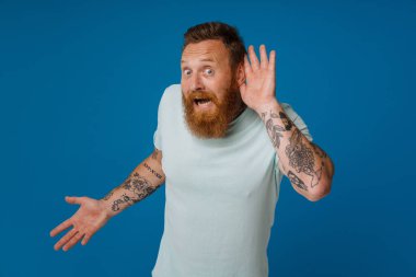 Ginger şok olmuş sakallı bir adam mavi arka planda izole edilmiş kameraya şaşırdığını ifade ediyor.