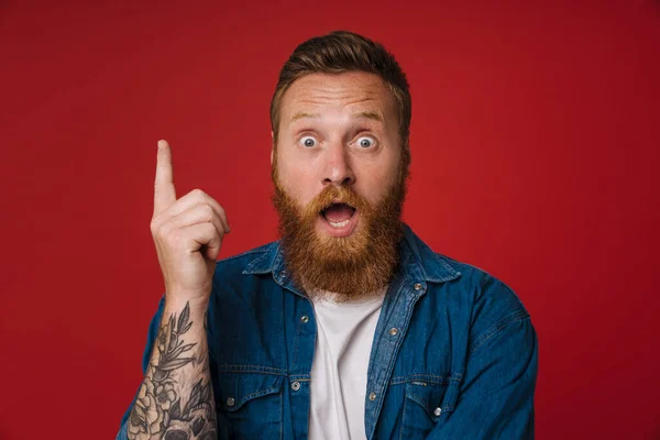 Ginger man expressing surprise and pointing finger upward isolated over red background