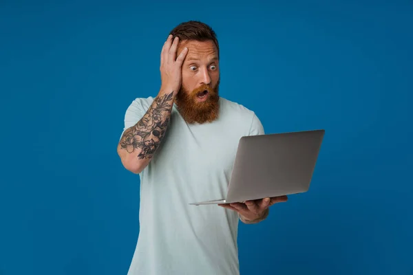 Imbir Wstrząśnięty Człowiek Wyrażając Zaskoczenie Podczas Pracy Laptopem Izolowane Niebieskim — Zdjęcie stockowe