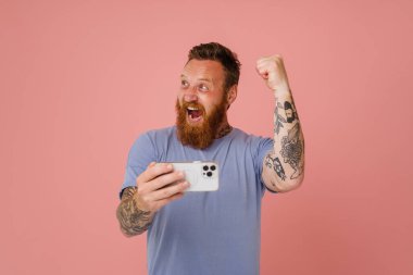 Kızıl beyaz adam pembe arka planda izole edilmiş cep telefonuyla oyun oynarken el kol hareketi yapıyor.