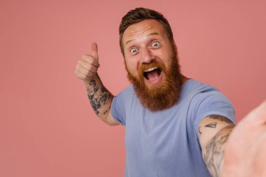 Kızıl heyecanlı bir adam pembe arka planda izole edilmiş selfie ve jest çekiyor.