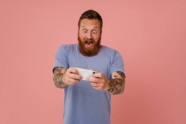 Kızıl beyaz adam pembe arka planda izole edilmiş cep telefonuyla oyun oynarken çığlık atıyor.