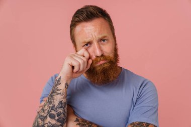 Ginger, sakallı üzgün bir adam pembe arka planda gözyaşlarını silerken ağlıyor.