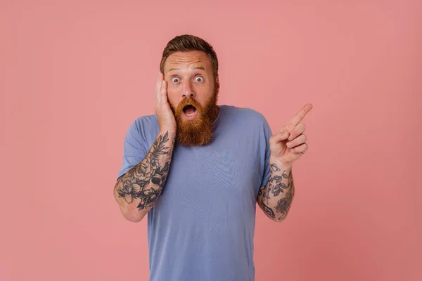 Ginger shocked man with beard screaming and pointing finger side isolated over pink background