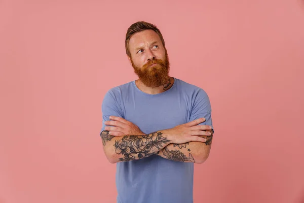 Ginger puzzled man with beard frowning and looking aside isolated over pink background