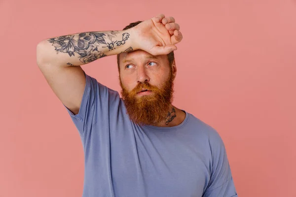 Ginger tired man with beard wiping his forehead and looking aside isolated over pink background