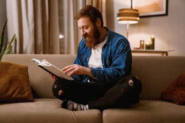 Zencefilli orta yaşlı adam rahat oturma odasında kanepede otururken kitap okuyor.
