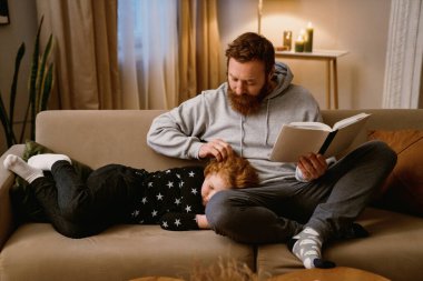 Babası kitap okurken küçük tatlı oğlu evdeki rahat oturma odasında dizlerinin üstünde uyuyor.