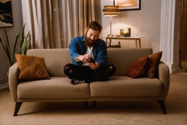 Heyecanlı sakallı adam evde rahat bir oturma odasında Lotus 'ta poz verirken cep telefonuyla oyun oynuyor.