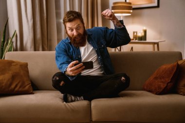 Heyecanlı sakallı adam cep telefonuyla oyun oynuyor ve Lotus poz verirken kazanan jestini sergiliyor.