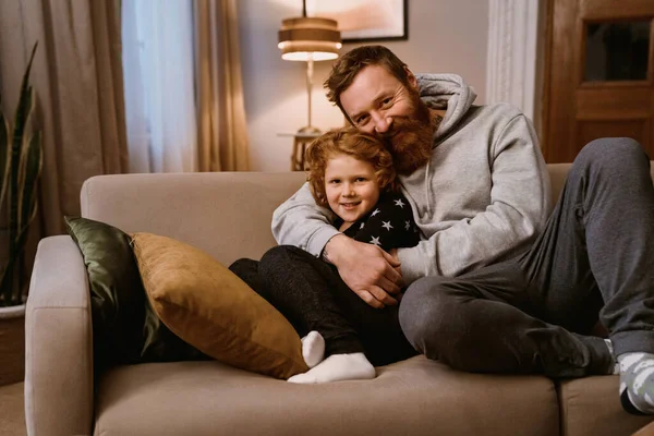 Ginger father and son hugging and smiling at camera while resting on sofa together in cozy living room at home