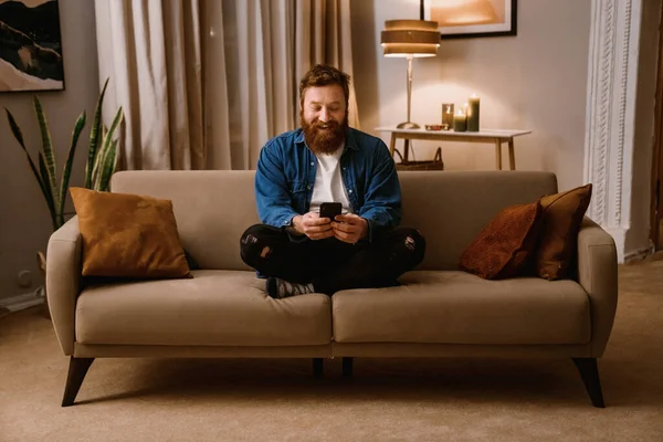 stock image Smiling redhead bearded man using mobile phone while sitting in lotus pose on sofa in cozy living room at home