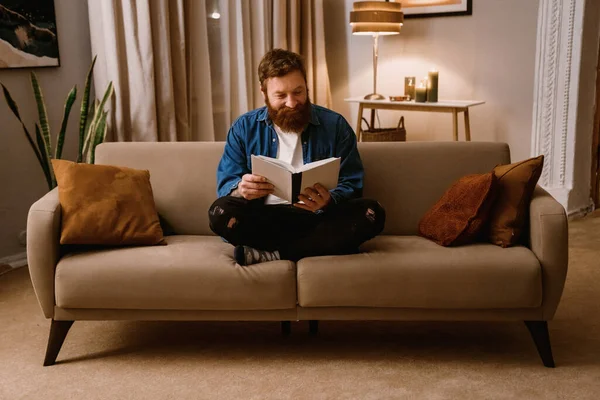 stock image Cheerful redhead bearded man reading book while sitting on sofa in cozy living room at home