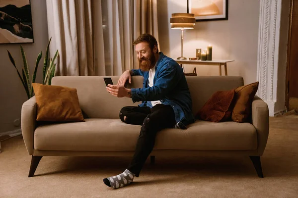 Stock image Smiling redhead bearded man using mobile phone while sitting on sofa in cozy living room at home