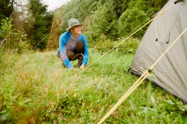 Yeşil ormanda yürüyüş yaparken panama giyen genç kızıl adam çadır kuruyor.