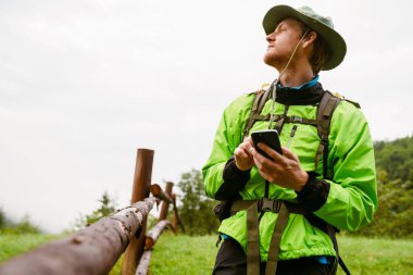 Panama giyen genç turist ahşap çitlerin yanında durup yeşil ormanda yürüyüş yaparken akıllı telefon kullanıyor.