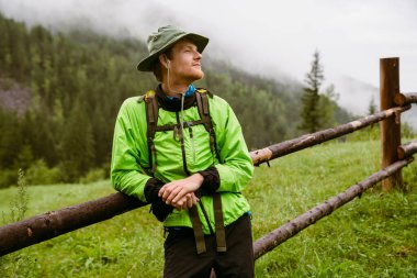Sırt çantalı genç turist ahşap çitlerin yanında duruyor ve dağlarda yürürken gözlerini kaçırıyor.