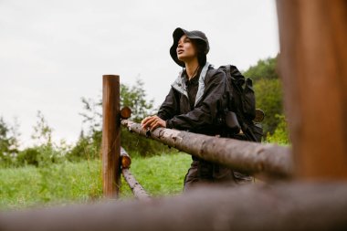 Yeşil ormanda yürüyüş yaparken tahta çitlerin yanında duran sırt çantalı Afrikalı genç bir kadın.