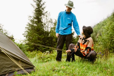 Yürüyüş ekipmanları giyen genç beyazlar dağ ormanlarında yürüyüş yaparken çadır kuruyorlar.