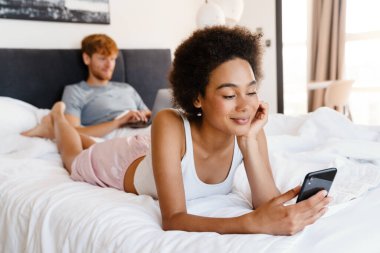 Genç, güzel, kıvırcık, gülümseyen, başını dik tutup telefonunu kullanan bir kadın. Evde dizüstü bilgisayarla çalışan erkek arkadaşıyla yatarken.