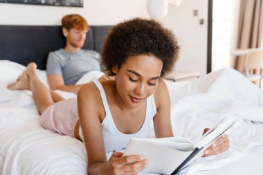 Genç ve güzel Afrikalı kıvırcık kadın evde dizüstü bilgisayarla çalışan erkek arkadaşıyla kitap okuyor.