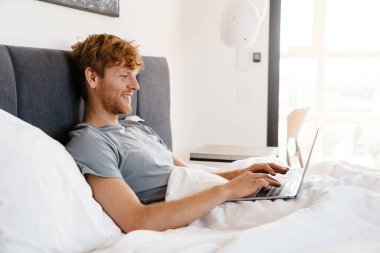 Genç, yakışıklı, gülümseyen, gri tişörtlü, laptopla çalışan, evde yatağında uzanan mutlu, kızıl saçlı adam.