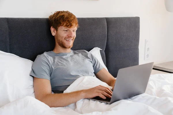 家でベッドの上に横たわっている間 ノートパソコンで作業しているグレーのTシャツで若いハンサムな笑顔の赤毛の男 — ストック写真
