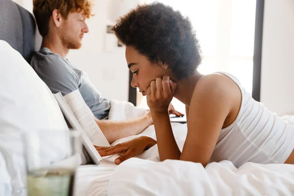 stock image Young beautiful african curly woman propping her head and reading book lying with her boyfriend who working with laptop on bed at home