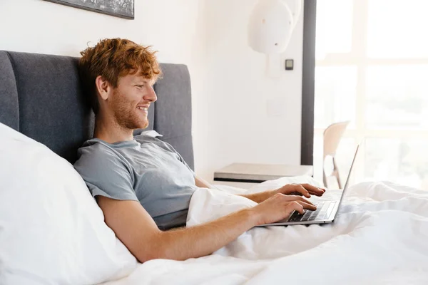 自宅でベッドの上に横たわっている間 ノートパソコンで作業しているグレーのTシャツで若いハンサムな笑顔幸せな赤毛の男 — ストック写真